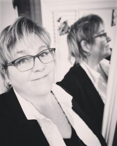  a black and white selfie of a smiling white nonbinary person with glasses and short hair, standing in front of a mirror that has their profile reflected in it."