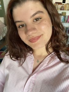 Brianna is white and has short auburn hair and is wearing a shirt with white and pink stripes. She is looking and smiling at the camera. Behind her there is a bookshelf and a green wall.