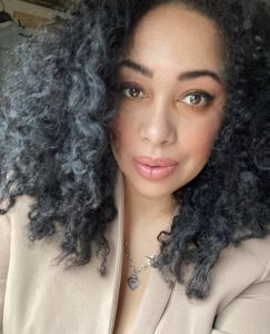 The image is of social media volunteer Kelsey White. Kelsey is a biracial, queer woman. Kelsey has black curly hair and brown eyes. Kelsey is looking into the camera while wearing a light beige blazer and silver heart necklace.