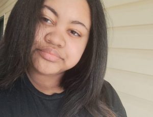 A photo of Amani, a black woman with black hair looking straight at the camera. She has a black shirt on.