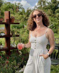 A woman with auburn, curly, shoulder-length hair stands outdoors in a garden on a sunny day. She is smiling while wearing tortoiseshell sunglasses and a white and gray pinstriped sleeveless jumpsuit with buttons down the front. She holds a mason jar filled with a pink drink in one hand and has the other hand in her pocket. Behind her is a wooden shelf with potted plants and flowers, with lush greenery and a blue sky in the background.