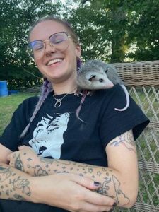 A photo of Elyse, a white Genderfluid person with tattoos and glasses, smiling. They are wearing their hair in two pastel purple braids, a black shirt, a silver necklace, and there is a small possum sitting on their shoulder.
The possum is called Mabel, she has a white face and tail with a grey body, grey ears, a pink nose, and black eyes.