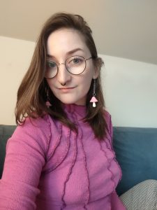 A selfie of Finn, a white nonbinary person with long brown hair who is smiling at the camera and wearing a magenta long sleeve shirt, pale pink mushroom earrings on long chains, black round glasses and a black circular septum ring.