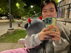 A photo of Nhi, a nonbinary Vietnamese-American sitting on a bench outside at night, wearing a large grey sweater and pink pants, their black hair tied back. They’re facing the camera, holding a stuffed animal in the crook of their arm and their friend’s phone in front of the bottom part of their face to take a picture of their friend off-screen.