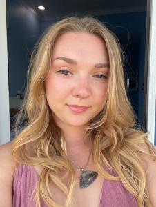 A nonbinary femme stands indoors near a doorway with sunlight illuminating their face. They have long, wavy blonde hair, fair skin, and blue eyes and are wearing a nose stud on the right side, a small septum ring, and a large triangular stone pendant on a silver chain necklace.